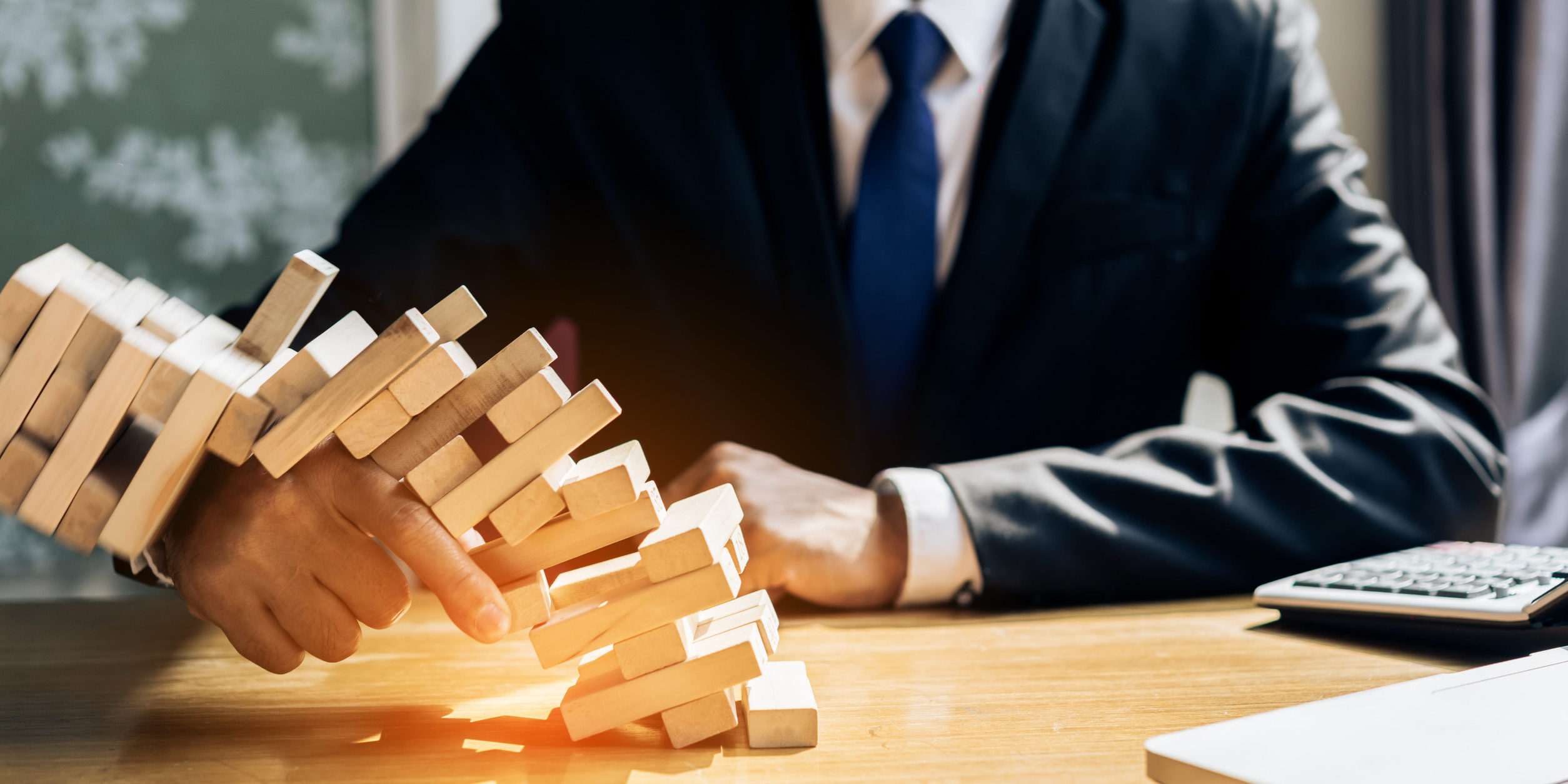 Person in a dark suit and blue tie plays a wooden Jenga tower on a office desk, hands steadying blocks.