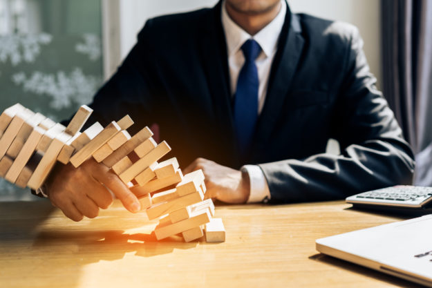 Person in a dark suit and blue tie plays a wooden Jenga tower on a office desk, hands steadying blocks.