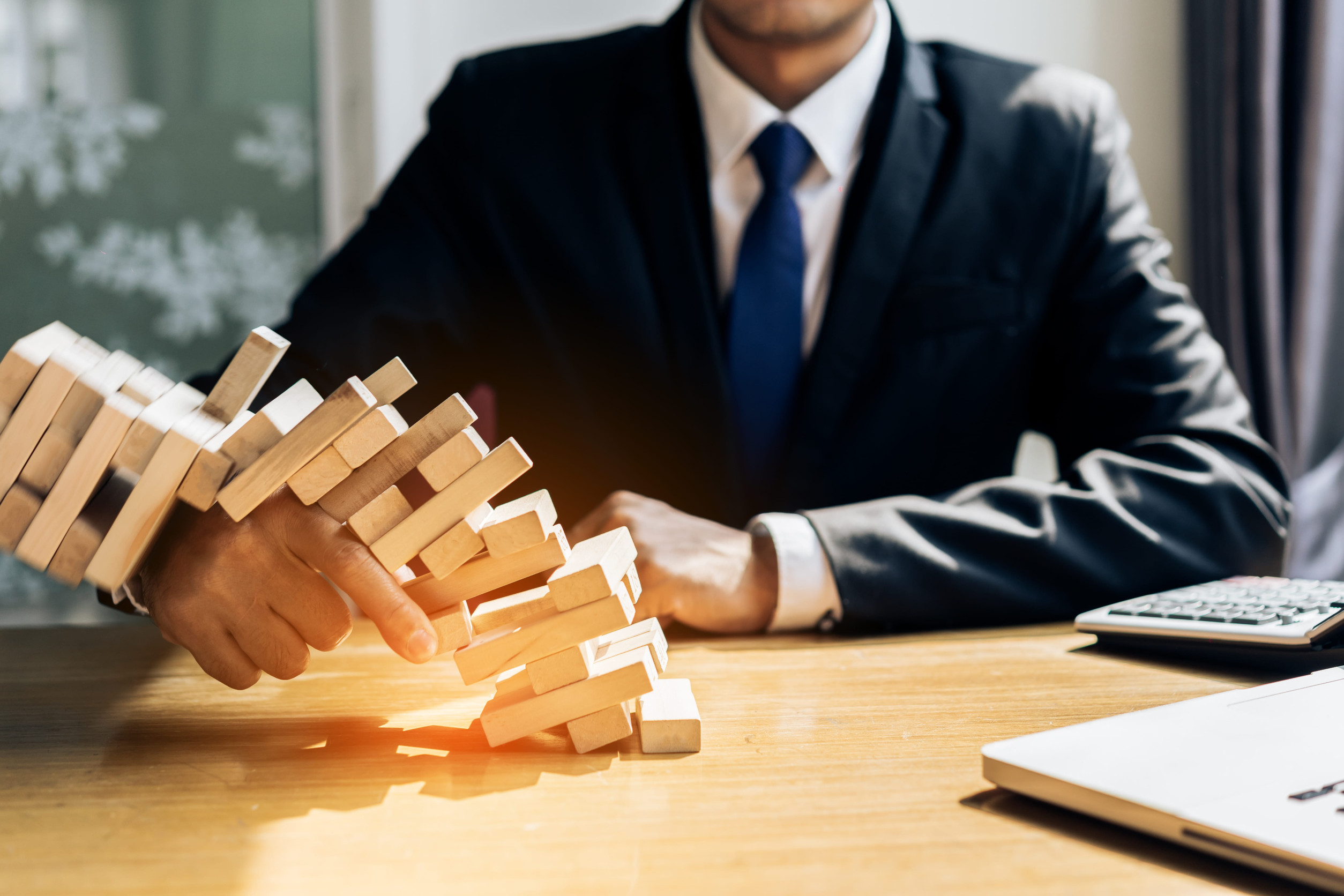 Person in a dark suit and blue tie plays a wooden Jenga tower on a office desk, hands steadying blocks.
