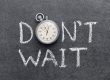 Stopwatch resting on a dark chalkboard with the words “DON'T WAIT” written in white chalk behind it.