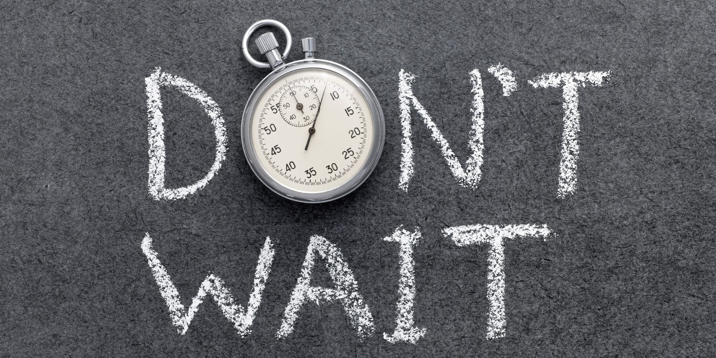 Stopwatch resting on a dark chalkboard with the words “DON'T WAIT” written in white chalk behind it.