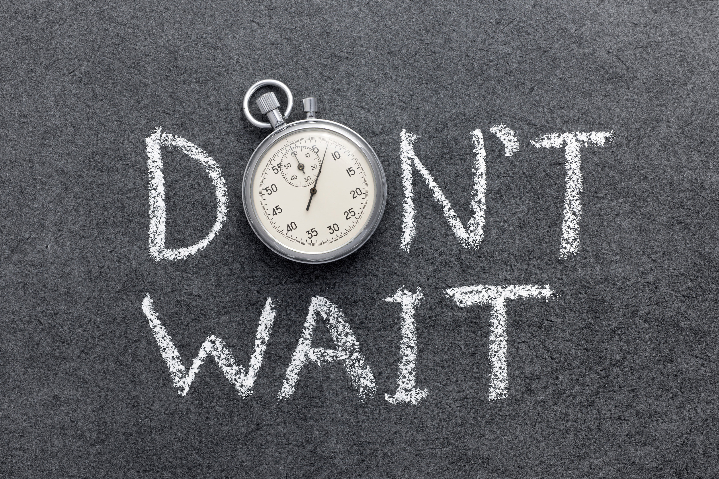 Stopwatch resting on a dark chalkboard with the words “DON'T WAIT” written in white chalk behind it.